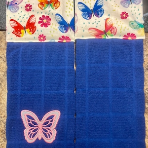 May include: Two blue kitchen towels with a white background featuring a colorful butterfly pattern. The towels have a pink embroidered butterfly on the bottom left corner.