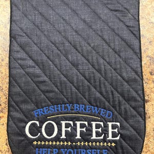 May include: A quilted, dark gray coffee pot holder with black trim. The holder features embroidered text in white and blue that reads "FRESHLY BREWED COFFEE HELP YOURSELF". The design includes a gold-colored line of stitching.