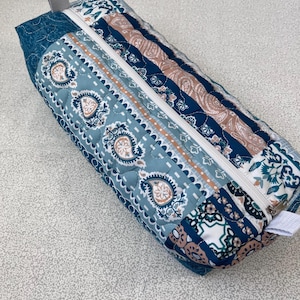 May include: A rectangular quilted pencil case with a zipper closure. The case features a patchwork design with various blue, teal, and tan paisley and floral patterns. A small gray fabric loop is attached to the end.