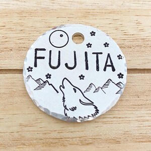 May include: Silver dog tag with a wolf howling at the moon, mountains, and stars in the background. The text "FUJITA" is engraved on the tag.