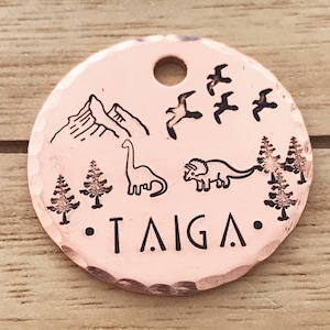 May include: A copper-colored round pet tag with a mountain, dinosaurs, trees, and birds engraved on it. The tag is personalized with the name "TAIGA".