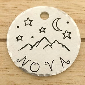 May include: A round, silver metal pet tag with a mountain range, stars, and a crescent moon design. The word "NOVA" is written below the mountain range. The tag has a hole at the top for attachment.