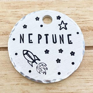 Op de afbeelding: Een ronde, zilverkleurige metalen tag met het woord "NEPTUNE" in zwarte hoofdletters. De tag heeft een zwarte raketillustratie, kleine zwarte bloem- en sterontwerpen en een gat aan de bovenkant voor bevestiging. De tag ligt op een houten oppervlak.