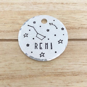 May include: Silver round pet tag featuring a constellation design and the name "REMI" engraved on it.