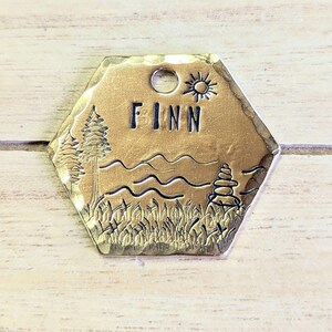 Op de afbeelding: Een zeshoekige hondenpenning van goudkleurig metaal met een zwart gegraveerd ontwerp. De penning toont een boom, bergen, golven, een zon en een stapel stenen. De naam "FINN" is in de penning gegraveerd.