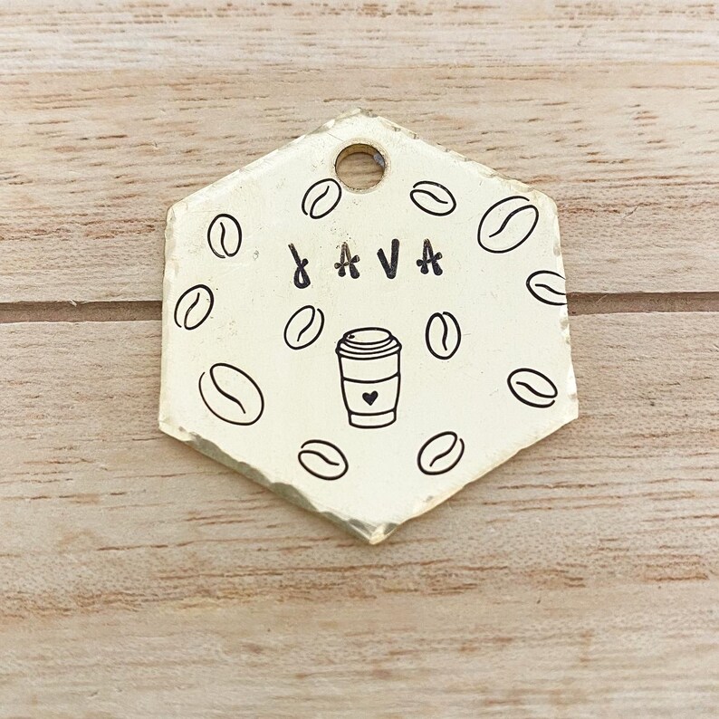 Op de afbeelding: Gouden zeshoekige hondenpenning met een koffiebeker en het woord "JAVA" erop gegraveerd. De penning is versierd met een patroon van koffiebonen.