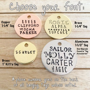 Op de afbeelding: Een selectie ronde metalen hondenhangers in verschillende maten en kleuren, met verschillende lettertypen en gegraveerde namen. De hangers worden gepresenteerd op een houten oppervlak. De tekst op de hangers luidt: "Raven Clifford Mocha Parker", "Rosie Alpha Nashville Cupcake", "Scarlet", "Sailor Molly Carter Magic".