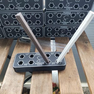 Puede incluir: Un sacapuntas triangular de pl&aacute;stico negro con dos ranuras para afilar l&aacute;pices. El sacapuntas tiene una etiqueta blanca que dice "Triangle Sharpener" y "Superior Cuts". Dos l&aacute;pices, uno marr&oacute;n y otro blanco, se est&aacute;n afilando en las ranuras.