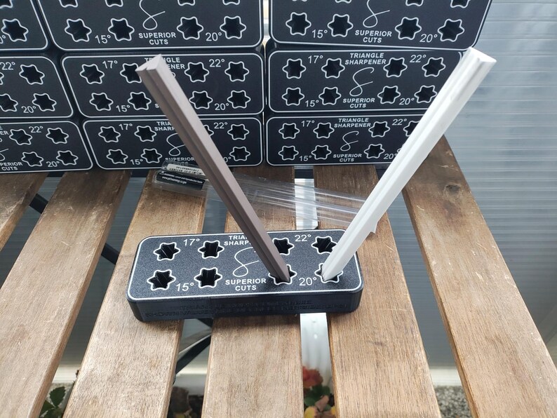 Puede incluir: Un sacapuntas triangular de pl&aacute;stico negro con dos ranuras para afilar l&aacute;pices. El sacapuntas tiene una etiqueta blanca que dice "Triangle Sharpener" y "Superior Cuts". Dos l&aacute;pices, uno marr&oacute;n y otro blanco, se est&aacute;n afilando en las ranuras.