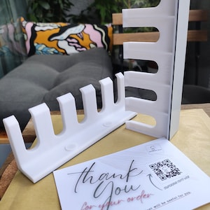 Puede incluir: Organizador de pared blanco de plástico con múltiples ranuras para guardar objetos. El organizador está montado en una superficie marrón. Una tarjeta de agradecimiento con un código QR está en primer plano.