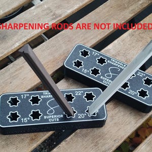 Puede incluir: Dos guías de afilado de plástico negro con ranuras en forma de estrella para varillas de afilado. Las guías están etiquetadas con diferentes ángulos: 15°, 17°, 20° y 22°. El texto "Triangle Sharpener" y "Superior Cuts" está impreso en las guías. El texto "Sharpening Rods Are Not Included" está impreso encima de las guías.