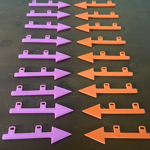 May include: Rows of purple and orange plastic arrows with a rectangular base and three small rectangular tabs. The arrows are arranged in two parallel columns on a dark surface.