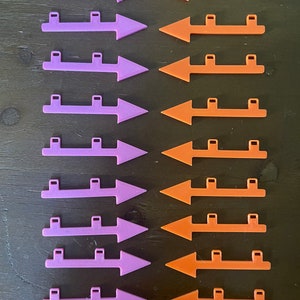 May include: Arrangement of purple and orange plastic arrows. Each arrow has a rectangular base with two small rectangular cutouts. The arrows are arranged in two columns, with the purple arrows pointing left and the orange arrows pointing right.