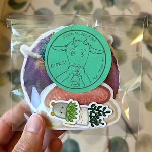 May include: A clear plastic bag containing a set of three stickers. The stickers feature a cartoon goat with the text "Omsies Mystery Pack" and "oops!" on a green circle, a salmon-colored sticker with gold glitter, and a sticker of a potted plant with a cartoon face.