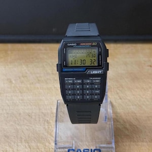 Puede incluir: Un reloj calculadora Casio Data Bank 80 negro con una pantalla digital que muestra la hora y la fecha. El reloj tiene una correa de goma negra y una cara de calculadora plateada con botones negros.