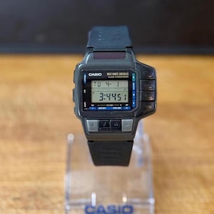 May include: A black Casio digital wristwatch with a black band. The watch face displays the time, date, and day of the week. The watch has multiple buttons on the side.