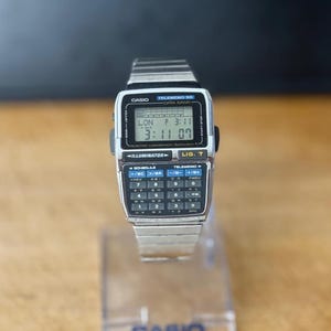 Puede incluir: Un reloj digital plateado Casio Telememo 50 con un teclado de calculadora y una pantalla digital que muestra la hora. El reloj tiene una correa plateada y el logotipo de Casio es visible. La pantalla muestra "LON P 3:11 3:11 07".