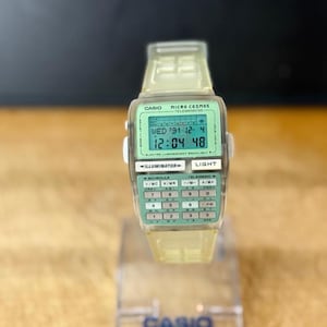 Puede incluir: Un reloj digital Casio Micro Cosmos Telememo 50 vintage. El reloj tiene una correa transparente y una esfera verde claro con un teclado de calculadora. La pantalla muestra la fecha y la hora. El reloj se muestra en un soporte transparente.