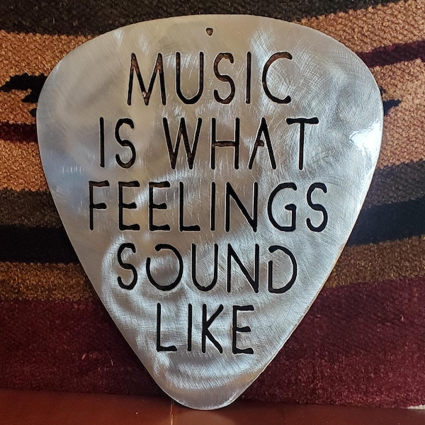 Könnte beinhalten: Ein silberner Gitarren-Plektrum aus Metall mit dem ausgeschnittenen Text "MUSIC IS WHAT FEELINGS SOUND LIKE".