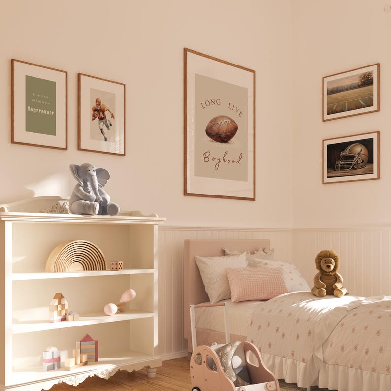 May include: A white bookshelf with three shelves, filled with toys. A gray elephant plush toy sits on top of the bookshelf. A brown teddy bear sits on a white bed with a pink and white comforter. There are four framed prints on the wall, one with the text "Long Live Boyhood" and a football illustration.