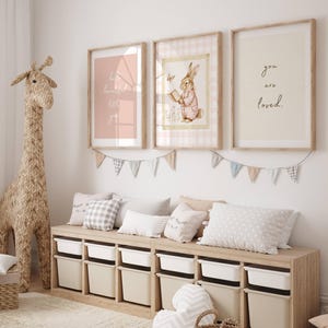 May include: A nursery room with a wooden bench with storage bins, three framed prints on the wall, and a woven giraffe toy. The prints feature a pink background with white text, a bunny reading a book, and the words "you are loved".