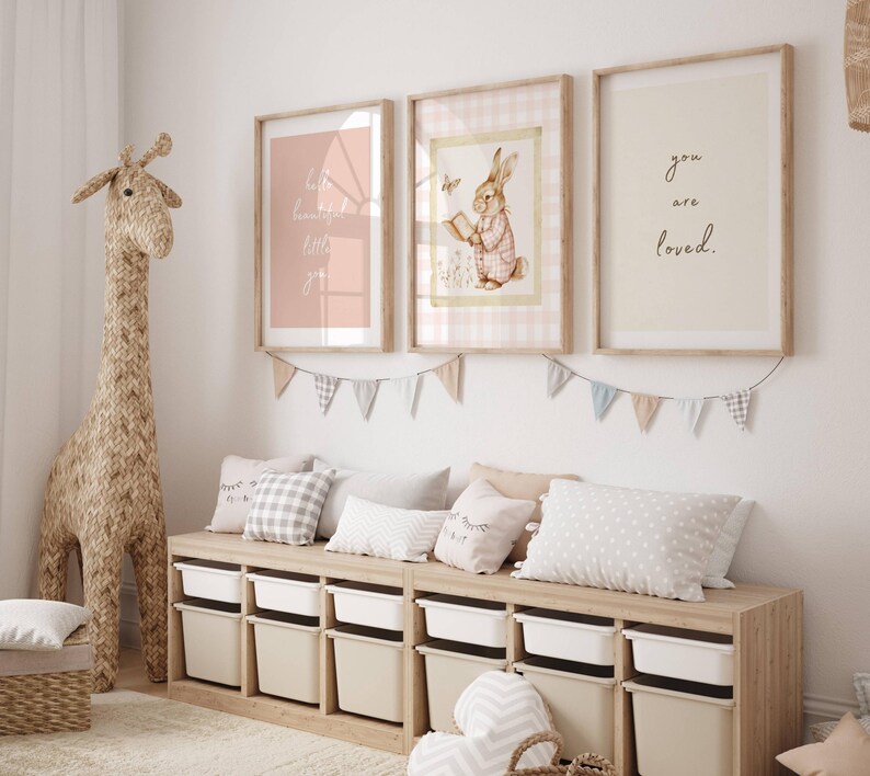 May include: A nursery room with a wooden bench with storage bins, three framed prints on the wall, and a woven giraffe toy. The prints feature a pink background with white text, a bunny reading a book, and the words "you are loved".