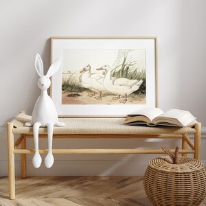 May include: A framed print featuring two white ducks standing near grass and water. The artwork is in a light wood frame, displayed above a woven bench with a white rabbit figurine and an open book. A woven apple-shaped basket sits nearby.
