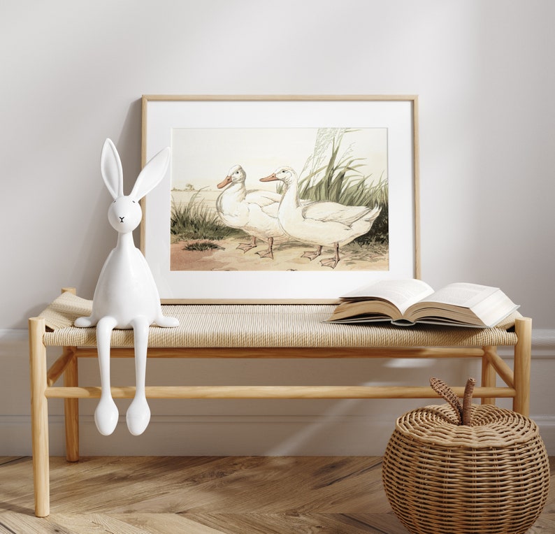 May include: A framed print featuring two white ducks standing near grass and water. The artwork is in a light wood frame, displayed above a woven bench with a white rabbit figurine and an open book. A woven apple-shaped basket sits nearby.