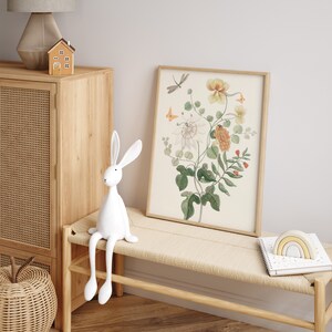 May include: A framed botanical print featuring a watercolor illustration of flowers, leaves, and insects. A white rabbit figurine sits on a woven bench. A wooden cabinet and a lamp are in the background.