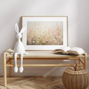 May include: A white ceramic rabbit figurine sits on a light wood bench with a woven seat. Behind the bench is a framed print of a watercolor painting of a field of white flowers with a soft pink and green background.