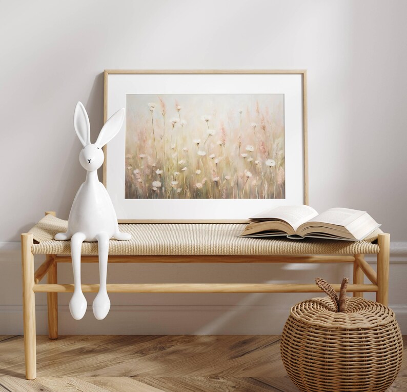 May include: A white ceramic rabbit figurine sits on a light wood bench with a woven seat. Behind the bench is a framed print of a watercolor painting of a field of white flowers with a soft pink and green background.