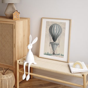 May include: A framed print of a vintage hot air balloon in shades of blue and white, displayed on a light wood bench. A white rabbit figurine sits on the bench, with a small wooden house and a lamp in the background. A woven basket is visible.