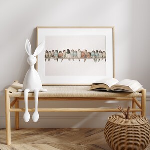 May include: A framed watercolor print of colorful birds perched on a branch. A white ceramic rabbit figurine sits on a wooden bench with a woven seat. An open book and a woven apple-shaped basket are also on the bench.