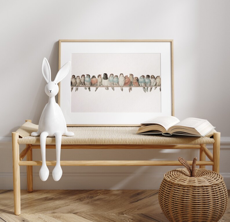 May include: A framed watercolor print of colorful birds perched on a branch. A white ceramic rabbit figurine sits on a wooden bench with a woven seat. An open book and a woven apple-shaped basket are also on the bench.