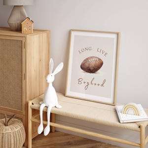 May include: A framed print with a watercolor illustration of a football and the text "Long Live Boyhood". The print is hanging on a wall above a wooden bench with a white rabbit figurine sitting on it.
