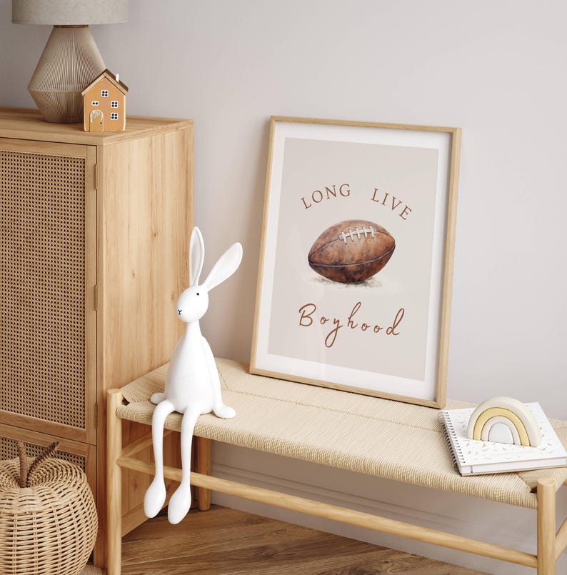 May include: A framed print with a watercolor illustration of a football and the text "Long Live Boyhood". The print is hanging on a wall above a wooden bench with a white rabbit figurine sitting on it.