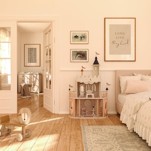 May include: A light wood toy castle with pink accents and a white roof. The castle has a mirror, a table, and a chair inside. The castle is on a light wood floor with a rug in front of it. There is a framed print on the wall that says "Long Live Boyhood".