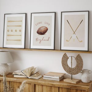 May include: Three framed prints with a beige background. The left print features a series of baseball bats and balls. The center print features a brown football with the text "Long Live Boyhood". The right print features two crossed golf clubs.