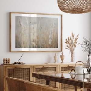 May include: A framed print of a watercolor painting depicting a field of tall grasses and flowers in shades of beige and white. The print is hanging on a white wall above a wooden cabinet with woven rattan doors.