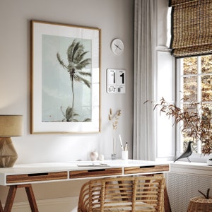 May include: A framed print of a palm tree with a white background. The print is hanging on a wall above a white desk with a wooden top. A wicker chair sits in front of the desk. The wall also has a clock and a calendar that reads "SAT 11".