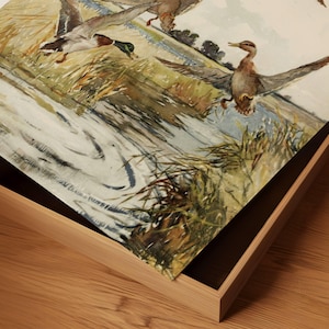 May include: A watercolor painting of three ducks flying over a pond. The ducks are brown and white with long necks and beaks. The pond is surrounded by green grass and reeds. The painting is framed in a wooden frame.