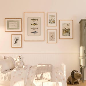 May include: A gallery wall featuring framed art prints with a fishing theme. Prints include illustrations of fish, ducks, and the text "Little Fisherman in the making" and "Long Live Big Fish". The frames are light brown, and the wall is painted a light pink.
