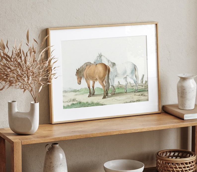 May include: Framed print featuring two horses in a field. One horse is brown, the other is white. The artwork is set in a light wood frame and displayed on a wooden shelf with decorative items.