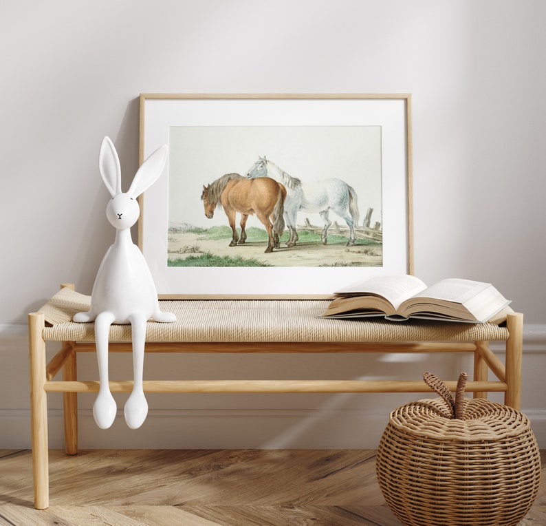 May include: A framed print featuring two horses, one brown and one white, standing in a field. The artwork is displayed on a light wooden bench with a white rabbit figurine and an open book. A woven apple-shaped basket sits nearby.