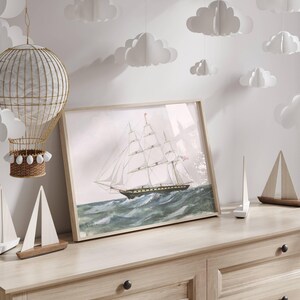 May include: A framed watercolor print of a tall ship sailing on a wavy sea. The artwork features a light wooden frame and is displayed on a light-colored wooden dresser. Decorative paper clouds hang from the ceiling, and small wooden sailboat models are on the dresser.