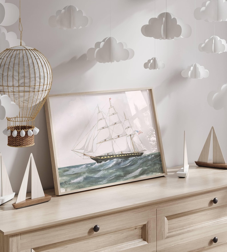 May include: A framed watercolor print of a tall ship sailing on a wavy sea. The artwork features a light wooden frame and is displayed on a light-colored wooden dresser. Decorative paper clouds hang from the ceiling, and small wooden sailboat models are on the dresser.