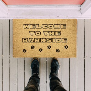 May include: A brown doormat with the text "Welcome to the Barkside" in black lettering. There are six paw prints in black below the text. A person's legs are visible at the bottom of the image, wearing black shoes and jeans.
