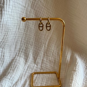 May include: A gold-colored earring display stand holding two gold hoop earrings. The earrings have a distinctive design. The stand is made of metal and has a rectangular base and a curved top with a small ball on the end.