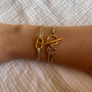 Peut inclure: Deux bracelets dorés. L'un présente un fermoir en forme d'ancre, l'autre un fermoir à bascule et un charme circulaire. Les deux sont portés sur un poignet, mettant en valeur leur design et leur couleur dorée.