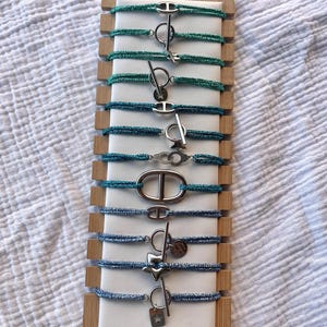 May include: A selection of bracelets with different clasp styles. The bracelets have a woven, glittery cord in teal and blue hues. The clasps are silver-toned, including toggle, oval, and charm designs. The display is presented on a wooden stand.